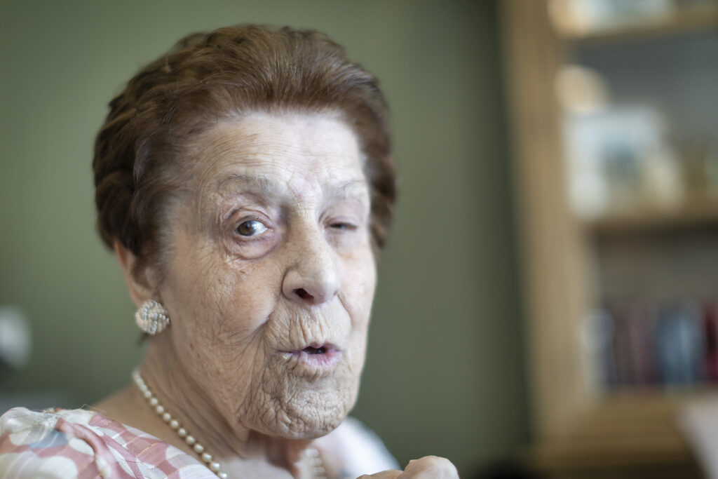 Een oudere vrouw met kort bruin haar, pareloorbellen en een ketting, zittend binnenshuis, knipogend en met een speelse blik, tegen een wazige groene en bruine achtergrond.