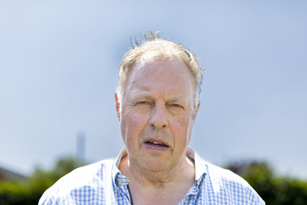 Een oudere man met dunner wordend haar en een geruit overhemd staat buiten op een zonnige dag en kijkt met een neutrale blik recht in de camera.