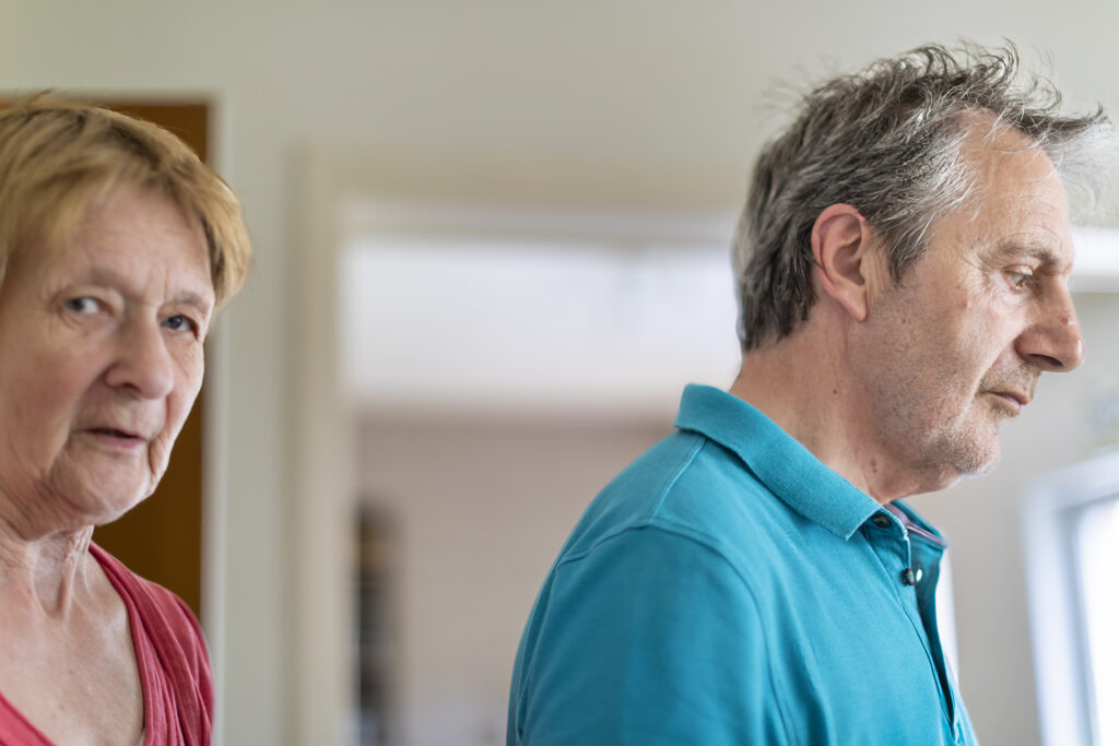 Een oudere vrouw in een rood shirt kijkt naar de camera, terwijl een oudere man in een blauwgroen shirt naast haar staat en met een ernstige blik de andere kant op kijkt, in een huiselijke omgeving.