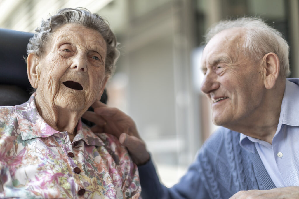 Een oudere vrouw in een bloemenblouse zit naast een oudere man in een blauwe trui. De man streelt zachtjes haar nek; ze glimlachen allebei hartelijk en genieten samen van een vrolijk moment.