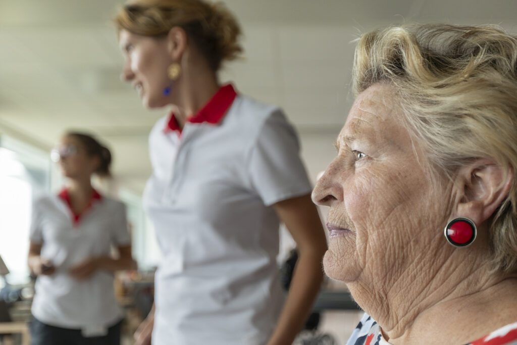 Een oudere vrouw met blond haar en rode oorbellen staat scherp in beeld, terwijl twee vrouwen in witte uniformen wazig op de achtergrond staan.