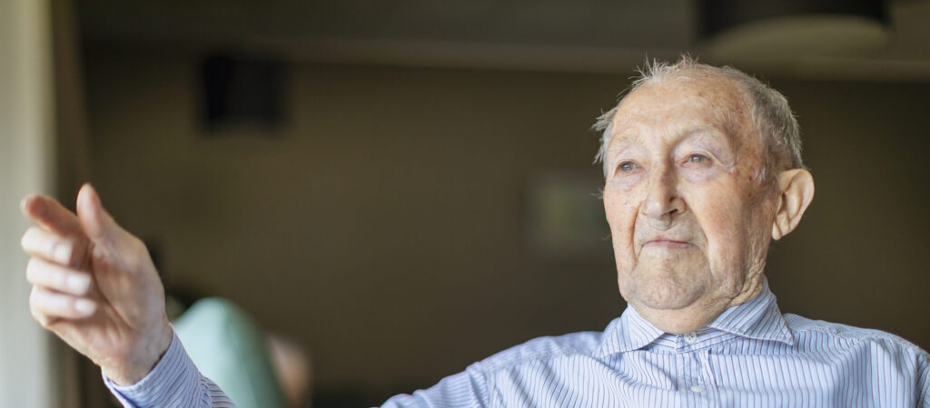 Een oudere man met kort grijs haar en een gestreept shirt zit binnenshuis, kijkt opzij en wijst met zijn vinger, met een neutrale uitdrukking op zijn gezicht.