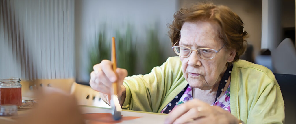 Een oudere vrouw met een bril en een groen vest schildert met een penseel op papier, terwijl ze geconcentreerd kijkt. Een binnenopname met een wazige achtergrond.