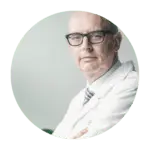 Een oudere man met een bril, een witte laboratoriumjas en een gestreepte stropdas staat met zijn armen over elkaar en kijkt naar de camera tegen een effen achtergrond.