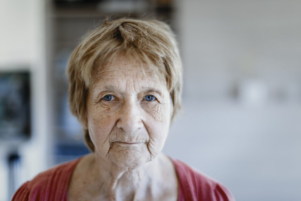 Een oudere vrouw met kort, lichtbruin haar en blauwe ogen kijkt recht in de camera; ze draagt een rode top. De achtergrond is zachtjes vervaagd, waardoor de aandacht volledig op haar peinzende blik wordt gericht.