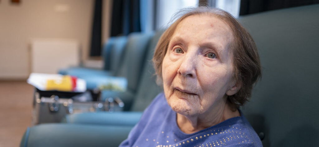 Een oudere vrouw met kort haar zit op een blauwe stoel; ze draagt een blauwe trui met kleine strasssteentjes. Ze kijkt iets voorbij de camera in een helder verlichte kamer, met wazige meubels en voorwerpen op de achtergrond.