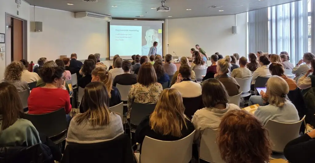 In een lichte vergaderzaal zit een groep mensen tegenover een spreker. Aan de voorkant wordt een dia met tekst en een afbeelding geprojecteerd. De meeste aanwezigen kijken naar voren en luisteren aandachtig.