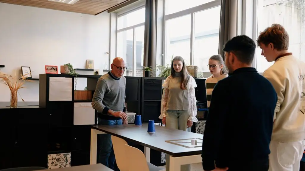 Een man demonstreert een bekerspel op een tafel, terwijl vier jongvolwassenen eromheen staan te kijken in een moderne, zonovergoten kantoorruimte.