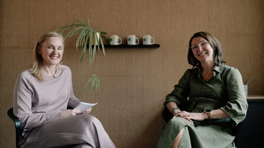 Twee vrouwen zitten glimlachend in stoelen in een warm verlichte kamer. Eén van hen houdt papieren vast, en op een plank op de achtergrond staan een plant en drie mokken.