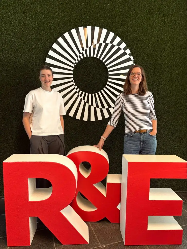 Twee vrouwen staan glimlachend achter grote rode letters R & E, voor een groene muur met een zwart-wit cirkelvormig kunstwerk.