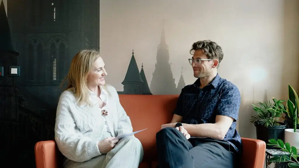 Twee mensen zitten op een oranje bank en voeren een gesprek. De vrouw houdt papieren vast en glimlacht, terwijl de man luistert. Naast hen staat een plant en op de achtergrond is een afbeelding te zien van hoge gebouwen in gotische stijl.