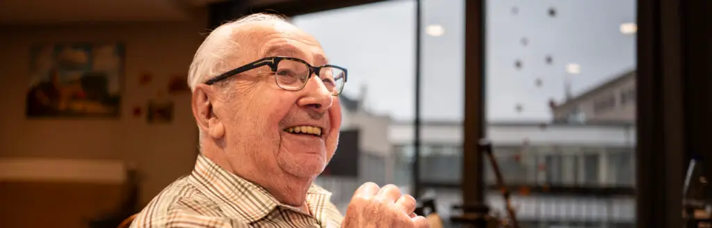 Een oudere man met een bril en een geruit overhemd zit op een bewolkte dag binnenshuis bij een groot raam en glimlacht.