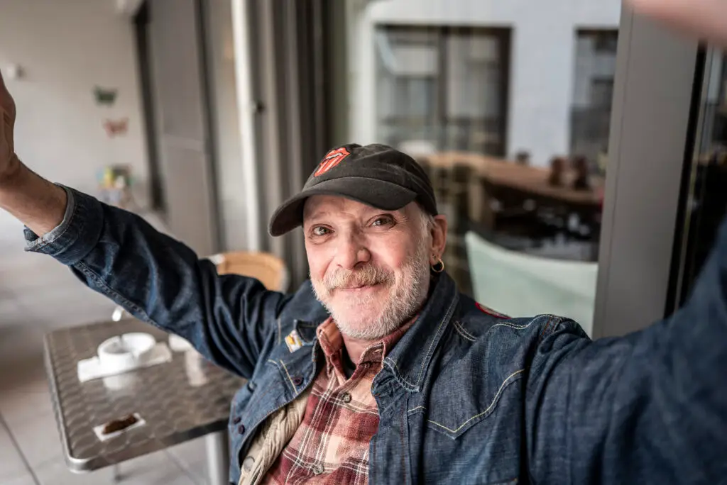 Een oudere man met een snor en baard, gekleed in een spijkerjasje, een geruit overhemd en een zwarte pet, zit met opgeheven armen te glimlachen aan een tafeltje op het terras van een café, met een kopje en schoteltje voor zich.
