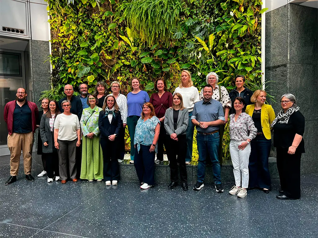 Een groep van twintig mensen poseert en lacht voor een grote groene binnenmuur die begroeid is met planten. De groep staat op een donkere, gespikkelde vloer in een modern gebouw met grijze stenen muren.