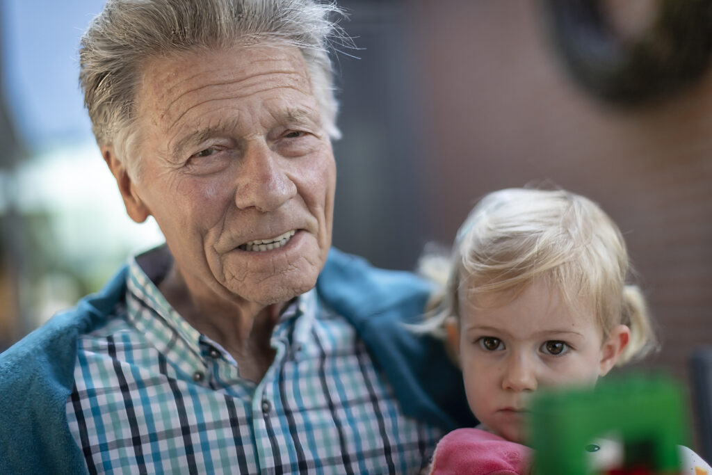 Een oudere man in een geruit overhemd glimlacht terwijl hij een jong blond meisje in het roze vasthoudt, dat er ernstig uitziet. De achtergrond is wazig, wat doet vermoeden dat de foto buiten is genomen.