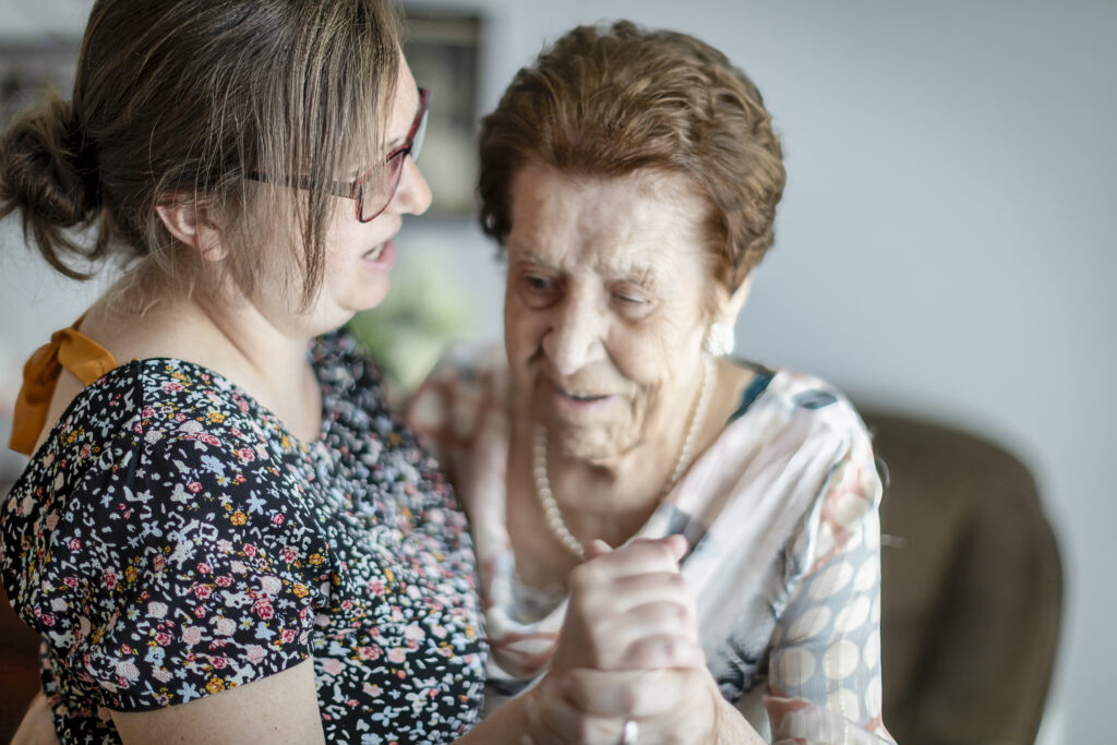 Een jongere vrouw met een bril en een bloemenjurk danst binnenshuis met een oudere vrouw in een gebloemde blouse; ze glimlachen allebei en houden elkaars hand vast.