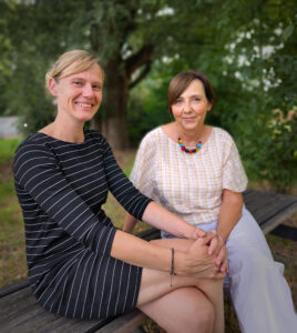 Twee vrouwen zitten dicht bij elkaar op een houten bankje in de buitenlucht en glimlachen naar de camera. De ene draagt een zwart gestreepte jurk en de andere een geruite blouse met een witte broek en een kleurrijke ketting van kraaltjes. Op de achtergrond is groen te zien.