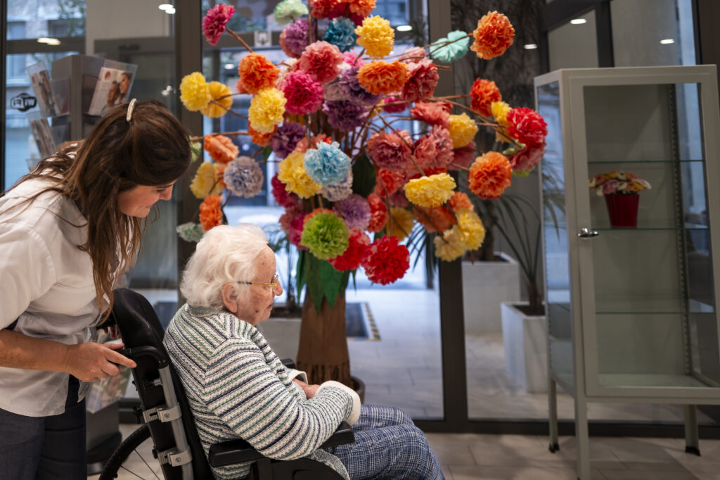 Een vrouw duwt een bejaarde in een rolstoel langs een kleurrijke kunstboom met felgekleurde papieren bloemen in een moderne binnenruimte.