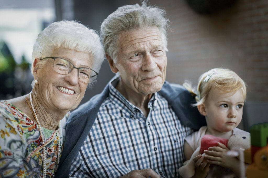 Een oudere vrouw en man glimlachen naast een jong meisje dat een stuk speelgoed vasthoudt. Ze staan dicht bij elkaar, buiten, en zien er gelukkig uit. De achtergrond is wazig, waardoor de aandacht vooral uitgaat naar hun gezichten en hun uitdrukkingen.