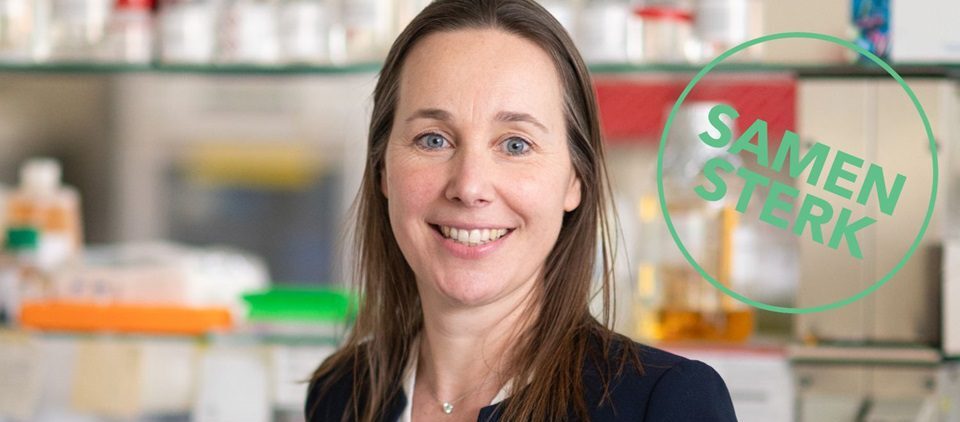 Een vrouw in een blazer glimlacht in een laboratorium. Op de achtergrond staan planken met flessen en laboratoriumbenodigdheden. Aan de rechterkant van de afbeelding staat een groene cirkel met de tekst ‘SAMEN STERK’.