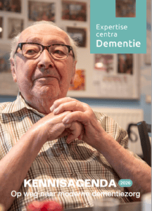 Een oudere man met een bril zit aan een tafel met zijn handen gevouwen. De tekst luidt: Expertisecentra Dementie en KENNISAGENDA 2026 – Op weg naar moderne dementiezorg...