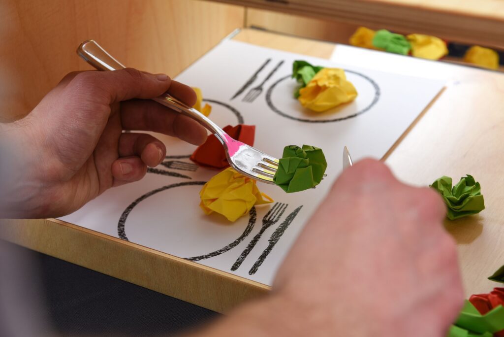 Iemand gebruikt een vork om een verfrommeld groen papieren balletje op te pakken van een placemat waarop een gedekte tafel is afgebeeld; dit dient als visueel hulpmiddel bij het omgaan met weerstand bij dementie.