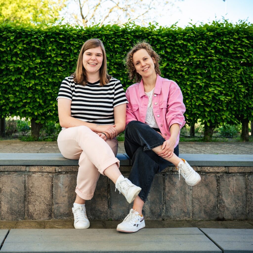Twee glimlachende vrouwen die op een laag stenen muurtje voor een heg zitten.